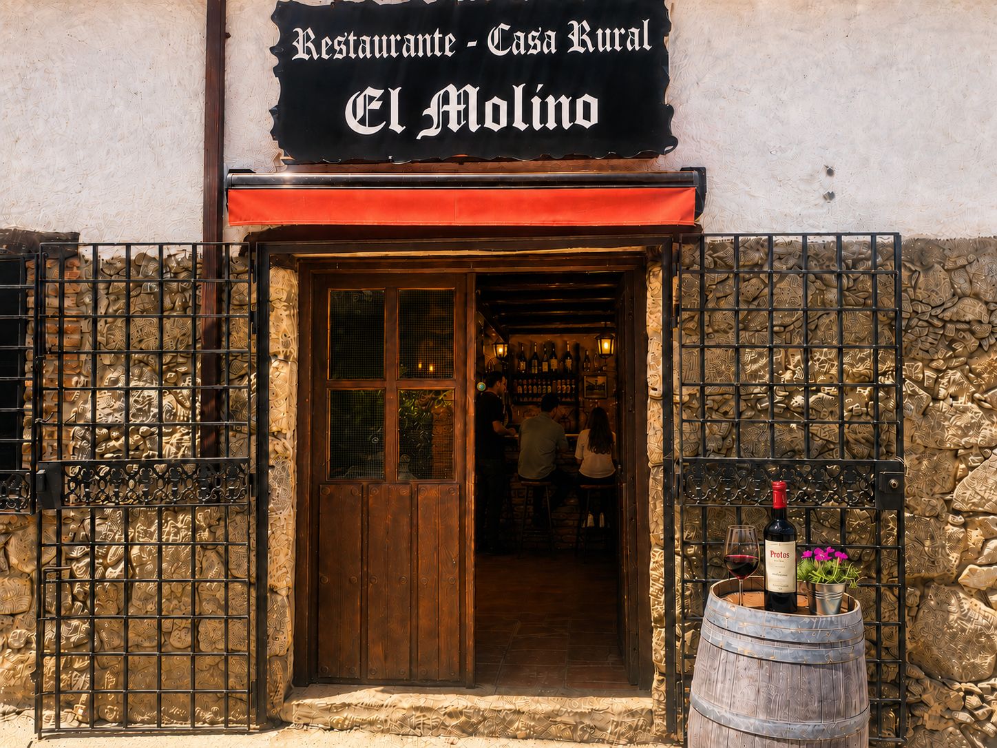 The stone-walled entrance of El Molino in Nuño Gómez (Toledo), its hand-painted sign above an open wooden door, with a barrel holding a bottle and glass of red wine